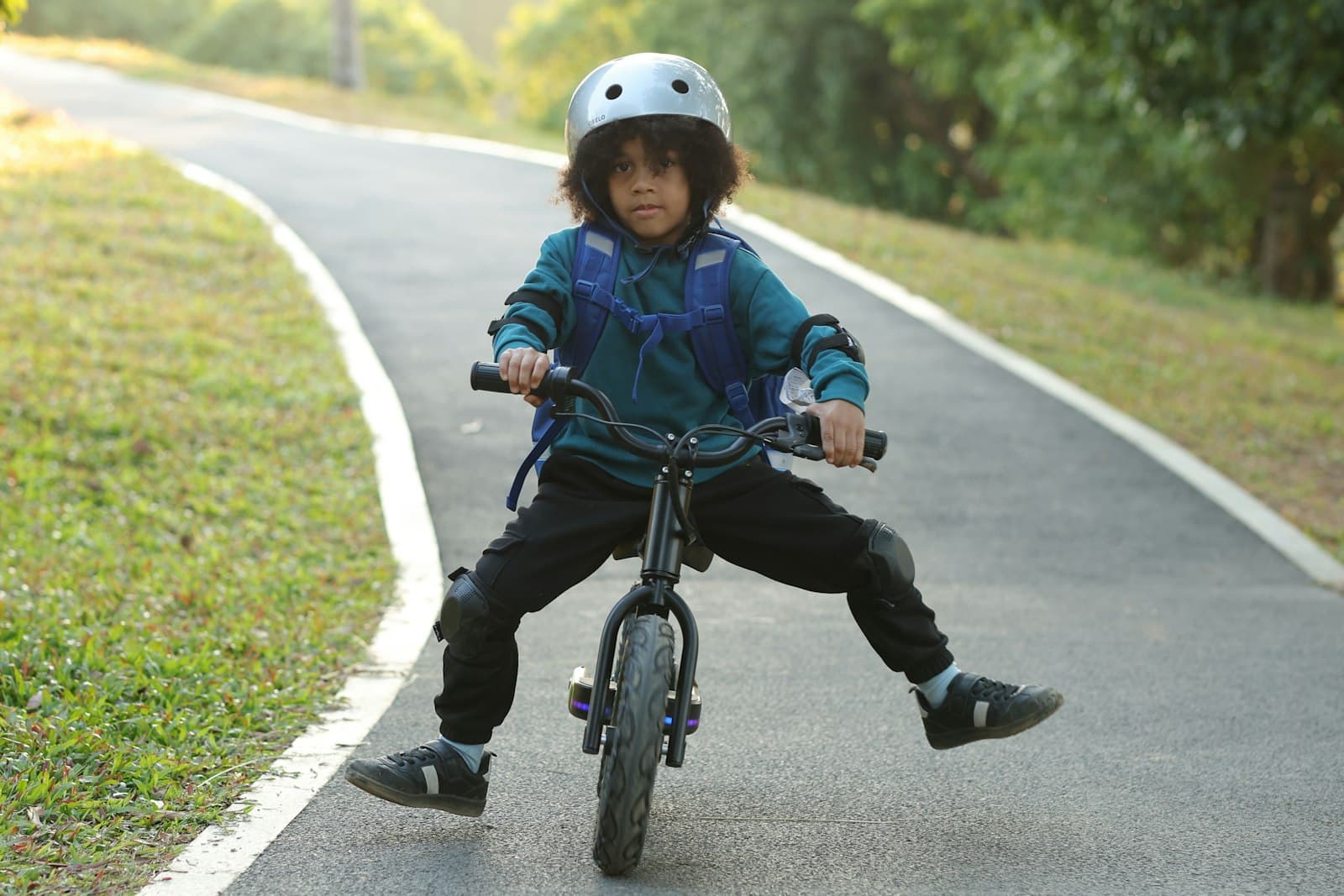 A young boy in a helmet and knee pads riding a balance bike along a park path