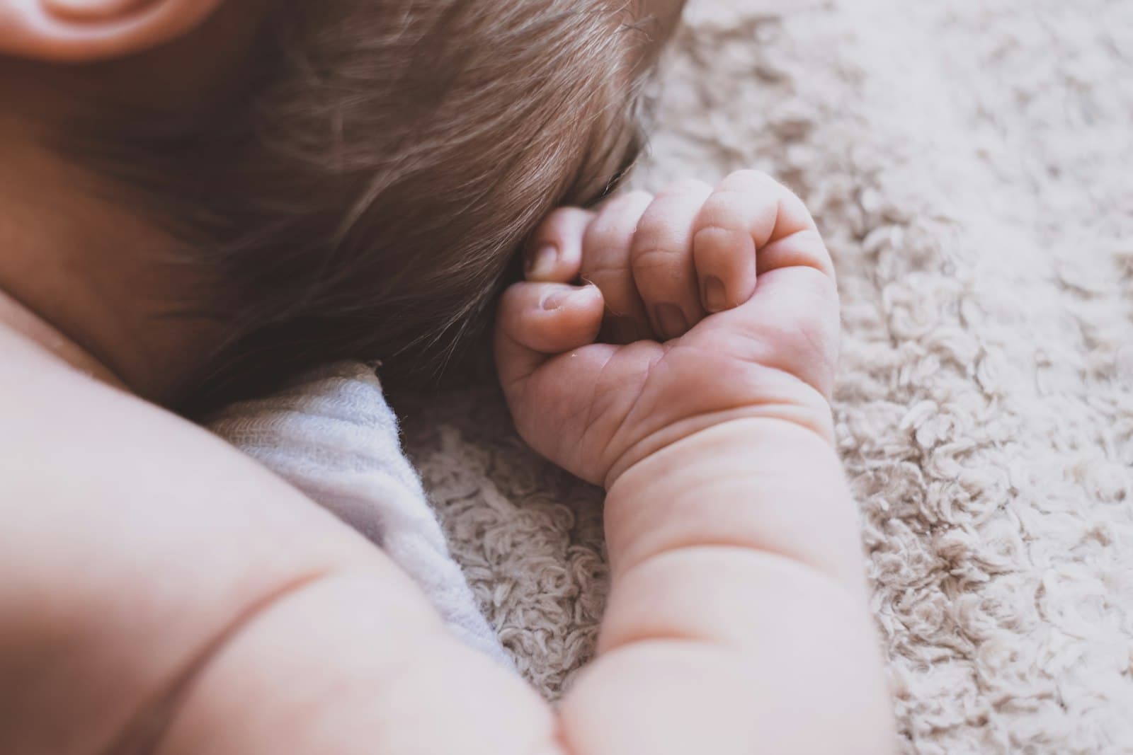 A newborn's hand resting by their head as they lie on a soft blanket
